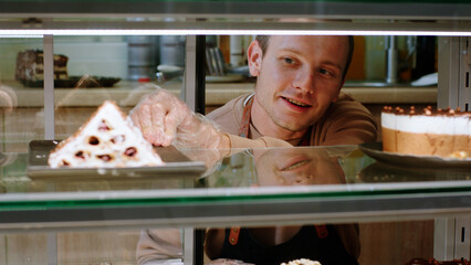 Closeup to the camera charismatic salesman in the bakery cafe carefully open the showcase fridge and arrange the deserts and add some fresh one he wearing protective mask in the pandemic of