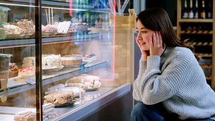 At the cozy coffee shop happy lady very hungry looking at the showcase fridge to choose her favourite dessert