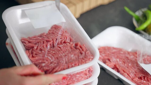 Fresh raw mixed beef and veal mince in white plastic containers, woman opening package with knife and removing food film from container, process of minced meat preparation at the kitchen for home