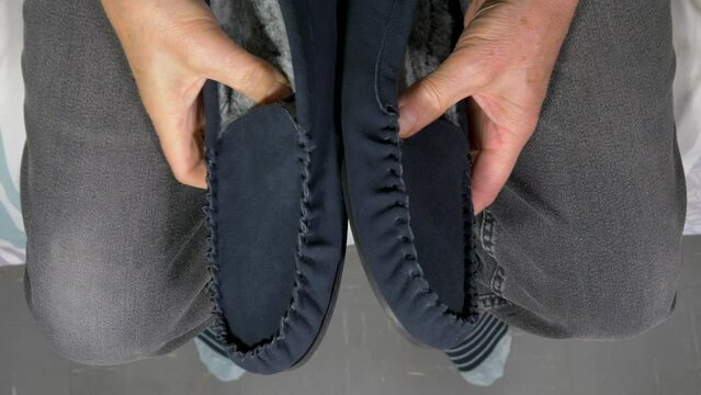 Closeup overhead shot of a senior man sitting on a bed, hands holding and checking a new pair of soft navy and gray, moccasin style slippers, with soft grey / gray lining.