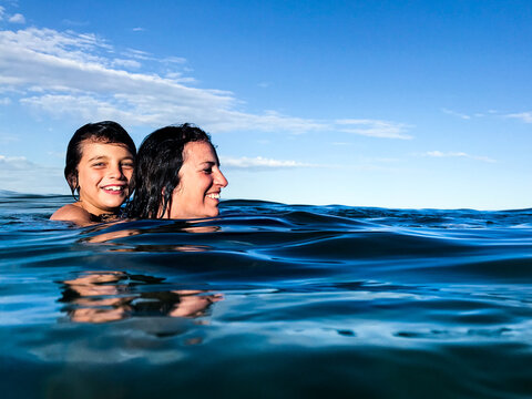 Mature woman and young boy on her back swimming through surface of calm blue ocean
