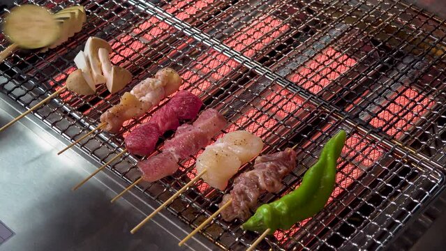 炭火の網の上で、鶏肉や野菜の串焼きをじっくりと焼き上げる手元のアップ。立ち上がる煙と肉汁が滴る様子、焼き鳥店や居酒屋の臨場感を伝える販促・広告用素材に。 | Grilled skewers (Yakitori) being cooked over charcoal on a wire rack with rising smoke.