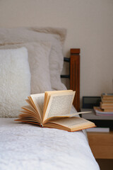 Old open book with yellow pages lies on a beige bed with pillows. Cozy fall mood in a room. Home education. Reading as a hobby. Mental health. Wellbeing, emotional support. Urban life. Indoor interior