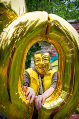 On a summer day, a girl in a yellow mask stands in nature and holds a yellow balloon in her hand.