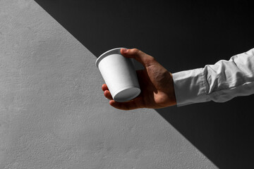 Man's hand holding blank coffee paper cup mockup, on concrete wall, with sharp natural sunlit...
