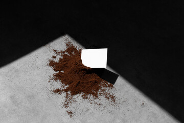 Blank business card in a pile of grounded coffee, on concrete background, with sharp natural sunlit...