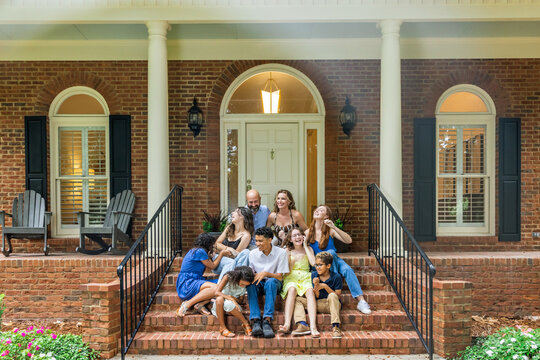 A large and blended and mixed happy family sitting on the front steps of their brick home in the suburbs