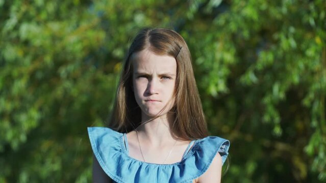 Sad teenage girl with long blonde hair raises head up to look into camera spending time by green trees in sunny summer park