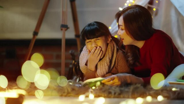 Pretty young mother reading a book to her daughter.happy Asian mom and daughter reading fantasy bedtime story book together at night time in kid tent at home before bed time with dim yellow light