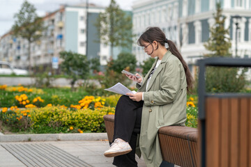 A girl sits on a bench on the street with documents in her hands and using her phone