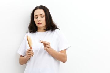 a funny, emotional, sad woman stands in a white T-shirt on a white background and looks sadly at the comb funny raising her eyebrows holding the fallen hair in her hand