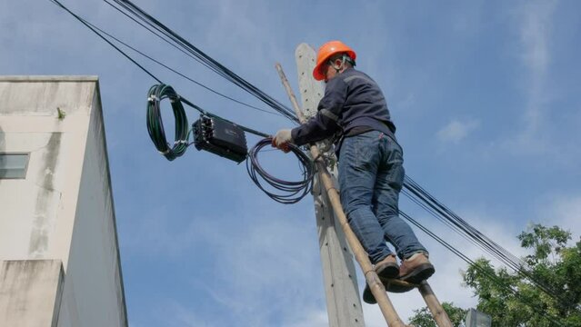 A telecoms worker is shown working from a utility pole ladder while wearing high visibility personal safety clothing, PPE, and a hard hat.