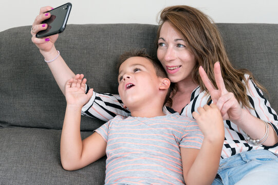 Cheerful Mother Holding Smart Phone Making Selfie With Her Little Son Showing Victory Sign. Mom And Kid Boy Sit On Couch Using Gadget Having Fun Taking Self-portrait Spend Time Together At Home.