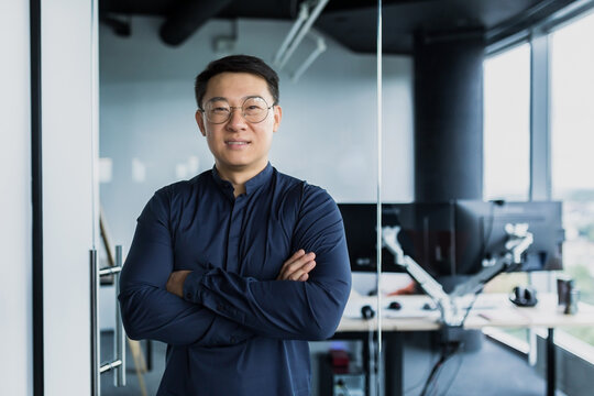 Portrait of successful Asian programmer, team leader company owner smiling and looking at camera with crossed arms, talented startup entrepreneur working inside IT office