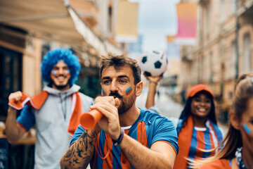 Soccer fan blowing vuvuzela while celebrating victory with group of friends on street.
