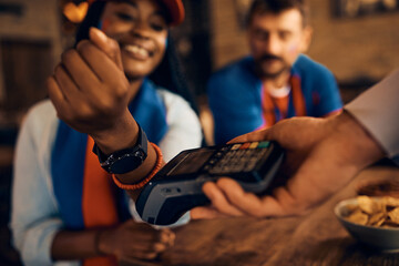 Close up of black sports fan using her smart watch while paying contactless in pub