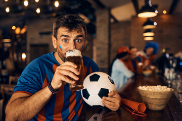Young soccer fan holding ball and drinking beer in pub.