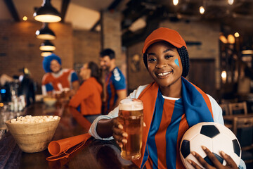 Happy black woman drinks beer while cheering for her team during soccer championship and looking at camera.