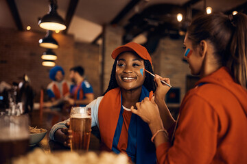Happy black woman drinks beer while getting her face painted before sports match in pub.
