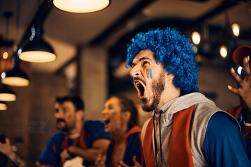 Excited sports fan screaming while watching match with friends in bar.