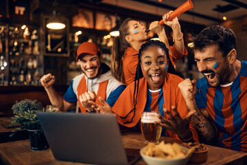 Group of excited sports fans celebrating their favorite team's victory while watching match on laptop in pub.