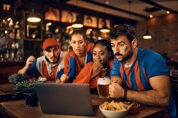 Young sports fan and his friends watching live broadcast of game on laptop in bar.