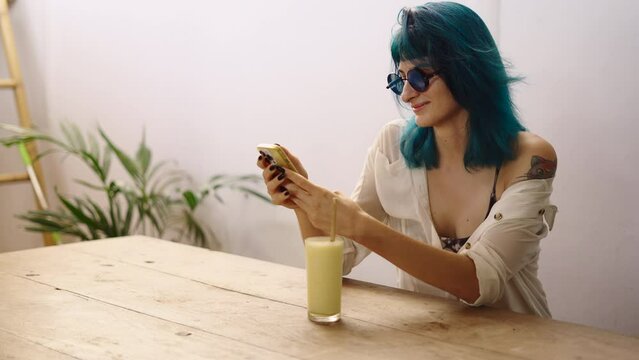 Alternative caucasian woman sits at table and text with smartphone while drinking pineapple juice 