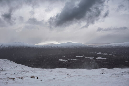 Epic Winter landscape image from mountain top in Scottish Highlands down towards Rannoch Moor during snow storm and spindrift off mountain top in high winds