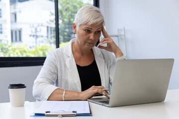 Business woman using digital table and working on laptop computer in modern home office.  Senior woman Serious working on laptop. Serious mature older adult woman watching on laptop working from home.