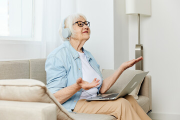 a happy modern old lady looks at the monitor holding a laptop on her lap talking through headphones smiling happily and gesticulating with her hands