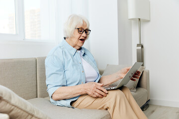 a lovely elderly woman is sitting in a cozy apartment and dreaming of messages on a laptop enthusiastically looking at the monitor while sitting on a beige sofa