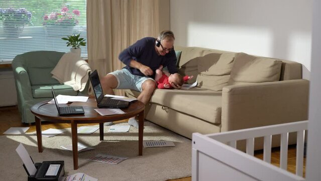 Middle-aged father with baby working from home and trying to calm the crying baby. A freelance single father with newborn makeing a video call and working with documents at home. Remote job, multi