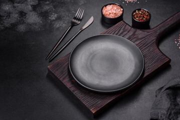An empty black plate on a dark concrete background with spices, herbs and cutlery
