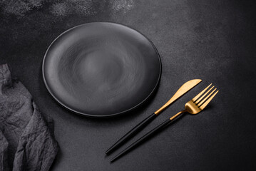 An empty black plate on a dark concrete background with spices, herbs and cutlery