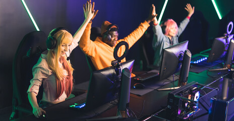 Three teenagers wearing colorful clothes and headsets using professional gaming setup celebrating...