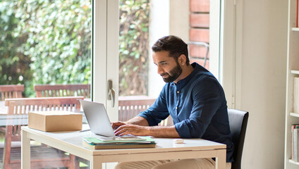 Indian ethnic business man, busy eastern male professional, serious businessman computing remote learning or distance working online typing on laptop using computer sitting at home office table.