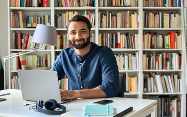 Happy indian professional business man, smiling teacher, student, business education coach sitting at work desk with laptop computer remote working, teaching or learning at home office, portrait.