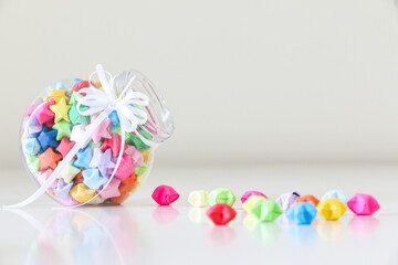 Handcrafted multicoloured origami lucky stars in a glass jar spilled onto white table. Lucky stars...