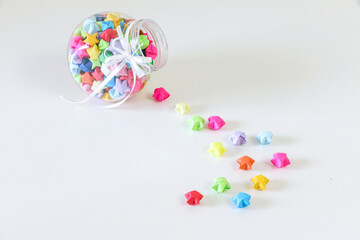 High angle shot of glass jar of colourful handmade origami paper lucky stars spilled onto white table. Lucky stars means a gift of love and luck.