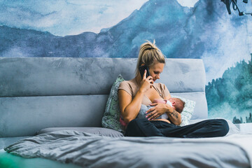 Young caucasian casually dressed mother sitting cross-legged on her grey bedspread, naturally feeding her little infant baby boy, and talking via smartphone. Modern bedroom interior. High quality