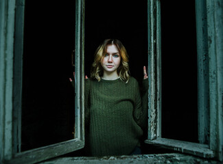A beautiful mysterious blonde young woman in a vintage green sweater stands at a broken window in the darkness of an old abandoned village house. Mysticism and horror.