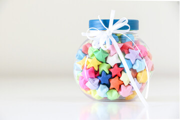Close up of handmade multicoloured origami paper lucky stars in a glass jar tied with a white ribbon on right side of frame. Lucky stars are a gift of love and luck.