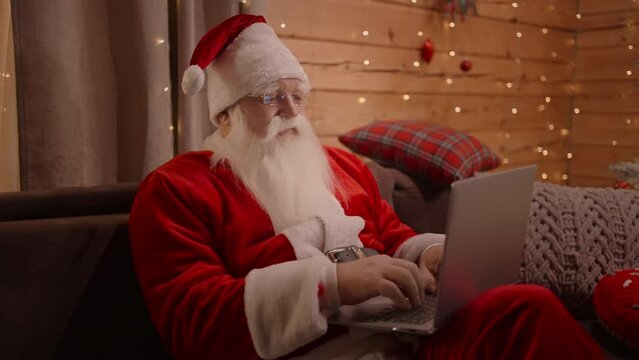 Portrait of Santa Klaus with a beard and glasses sitting with a laptop on Christmas Eve ordering gifts in the online store