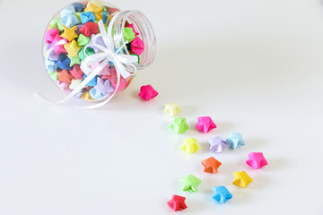 High angle shot of multicoloured handcrafted origami paper lucky stars in a glass jar spilled onto white table. Lucky stars wishing you love and luck.