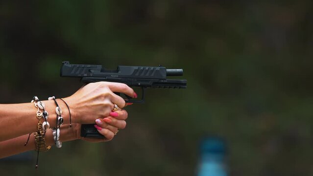 Loaded black gun held by unrecognizable caucasian woman with long red nails and lots of shell bracelets on wrists. Outdoor closeup slow motion shot of shell-case flying out. High quality 4k footage