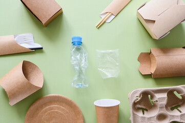 Eco-friendly paper utensils and packaging lie around plastic trash on a green background. View from...