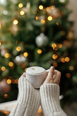 Hands holding warm cup of coffee on background of christmas tree with lights. Cozy home,...