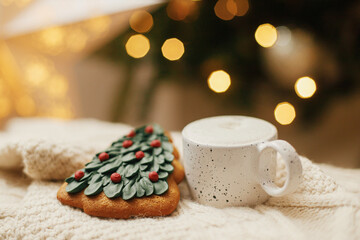 Atmospheric winter hygge. Warm cup of coffee and christmas tree gingerbread cookie on knitted sweater on background of christmas tree with lights. Christmas banner. Cozy holidays