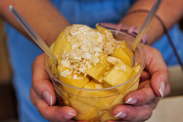 Close-up to hand holding a transparent cup filled with mango flavored ice cream and toppings.