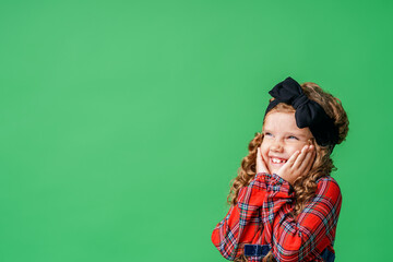 little girl of 7 years old with curly hair and a bow on her head, in a plaid dress, standing and smiling happily on a green background. Close-up. the model is having fun and feels stunned, amazed.